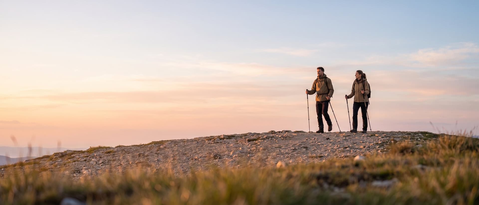 Two hikers with daypacks and trekking poles walking along a mountain ridge at sunrise under a wide sky.